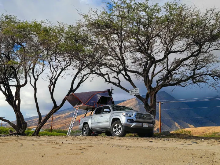 RV rental adventure: Silver truck with a rooftop tent pitched on sandy ground, framed by trees and distant mountains.. Toyota Tacoma 2017