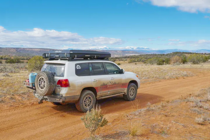 An off-road SUV with a rooftop tent, ideal for an RV rental adventure, drives on a dirt road through a vast desert landscape with snow-capped mountains.. Toyota Land Cruiser 2016