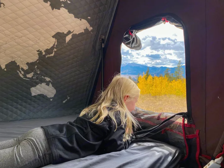 Inside an RV rental, a child with blonde hair watches golden fall trees & mountains from a map-lined tent.. Toyota Land Cruiser 2016