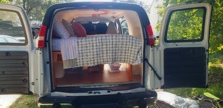 Converted van RV rental interior: cozy bed with pillows, patterned bedding, & under-bed storage, with open rear doors.. Chevrolet Express 2018