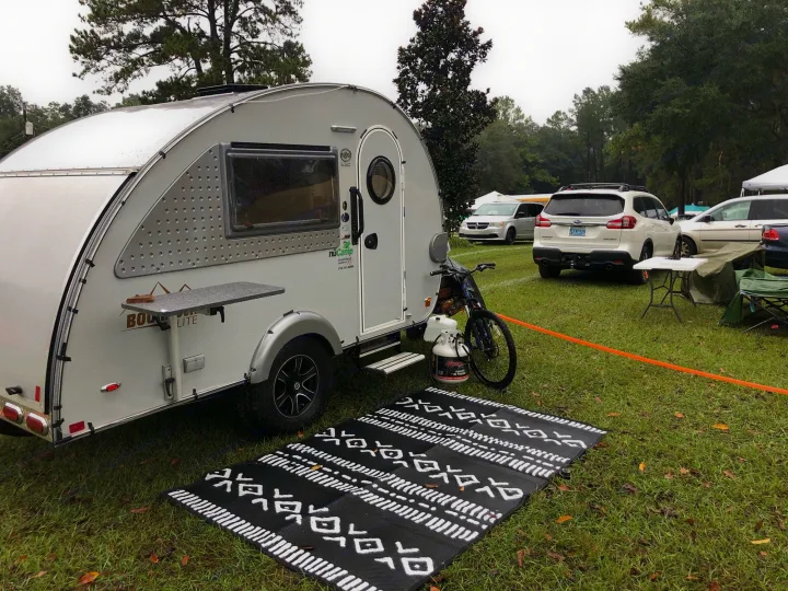 NuCamp BOOM Lite teardrop RV rental parked on a grassy campsite, complete with outdoor mats & a bike.. T@B T@B 320 OffRoad 2020