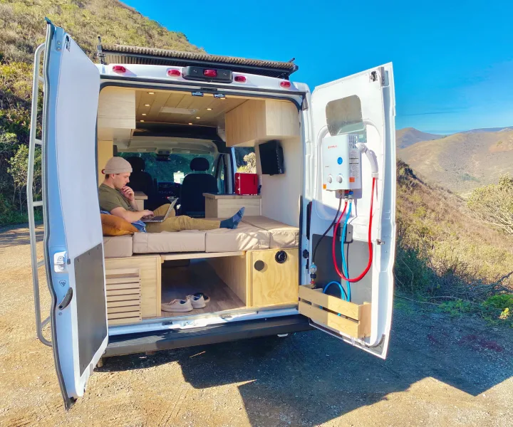 Remote work in style with this RV rental! Man uses laptop in a camper van with a beautiful view & outdoor shower.. ram ProMaster 2021