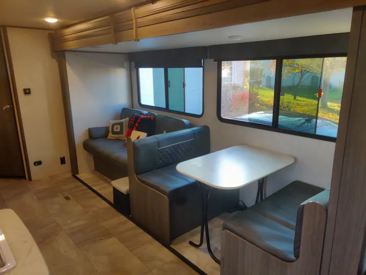 An RV rental's cozy interior shows a dark gray sofa, dinette, patterned floor, and bright windows with an autumn view.. Dutchmen Kodiak 2021