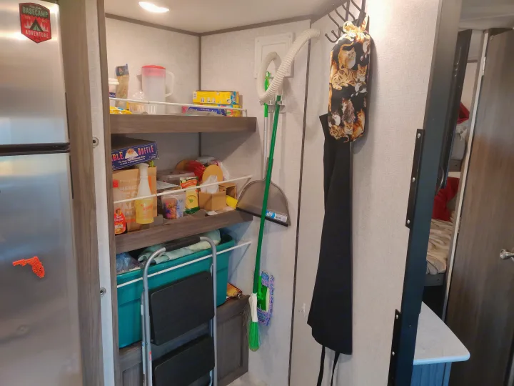 An organized RV rental interior, showing a kitchen pantry with food, cleaning tools, and a compact step ladder.. Dutchmen Kodiak 2021