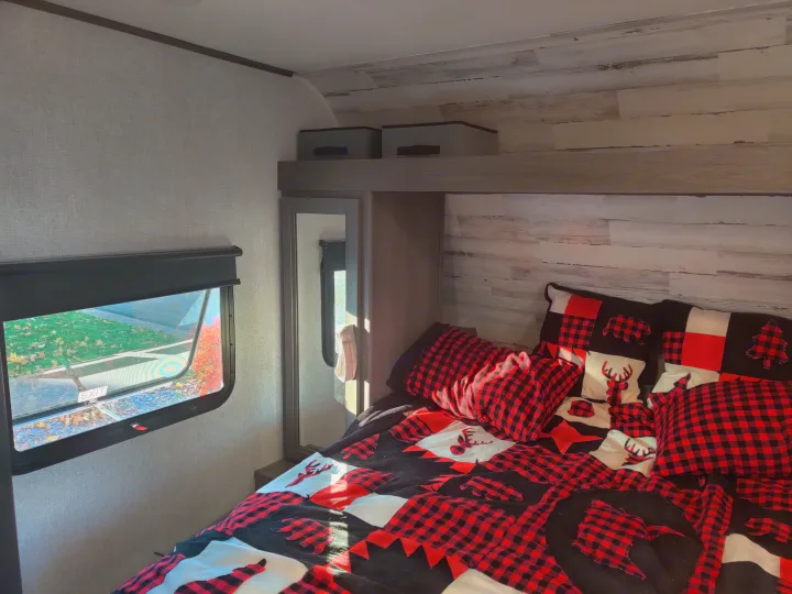 Cozy RV rental bedroom with red & black buffalo plaid bedding. Features deer, bear, tree designs, a window with outdoor view, and storage.. Dutchmen Kodiak 2021