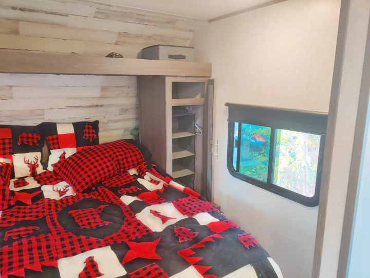 Cozy RV rental bedroom featuring buffalo plaid bedding, an upper bunk, and a window view of outdoor greenery.. Dutchmen Kodiak 2021