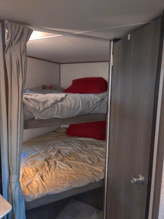 An RV rental interior shows a compact sleeping area with a bed, red pillow, and an overhead cabinet with a reading light.. Dutchmen Kodiak 2021