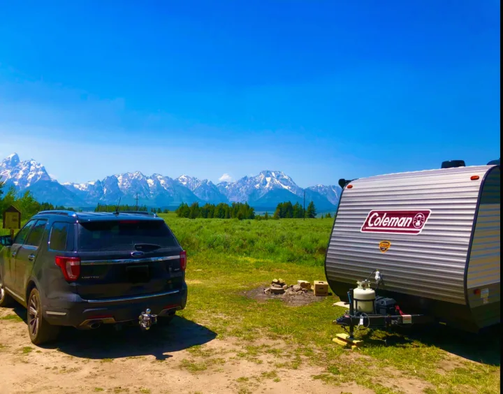 Camping scene: dark SUV and Coleman RV rental in a green meadow. Snow-capped mountains under a clear blue sky complete the scenic view.. Coleman Lantern LT 17B 2021