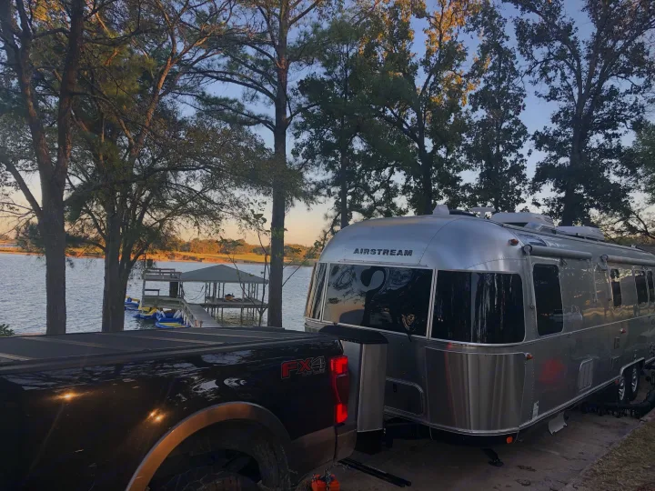 Black Ford FX4 pickup hitched to a shiny Airstream RV parked by a serene lake with a dock, ideal for rv rental.. Airstream Classic 2021