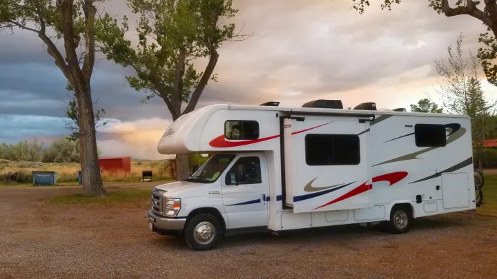 RV rental parked at a scenic campground under a dramatic, sunset-tinged sky, rain visible in the distance. Ideal for nature travel.. Forest River Sunseeker 2019