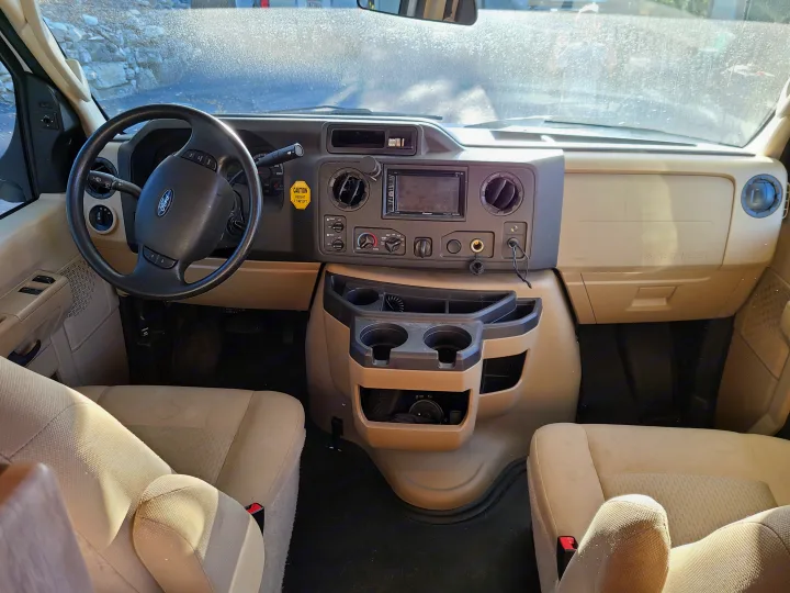 Interior view of an RV rental cockpit, featuring a Ford steering wheel, dashboard controls, and beige seats. A height caution sticker is visible.. Forest River Sunseeker 2019