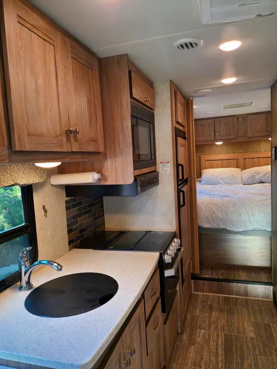 Spacious RV rental interior with light wood kitchen cabinets, sink, stovetop, microwave, and a cozy bedroom beyond.. Forest River Sunseeker 2019