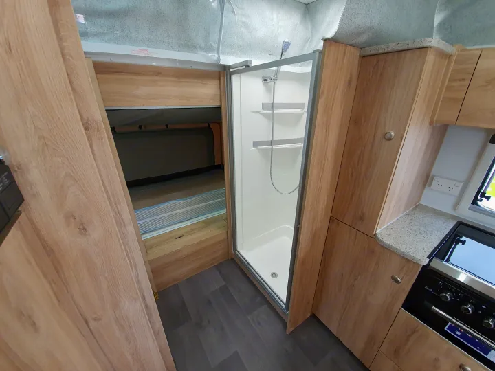 Inside an RV rental: view of compact shower, kitchen area with stove/oven, and a folded bunk bed, all with light wood cabinetry.. Golf Maxxi 501-1 2021