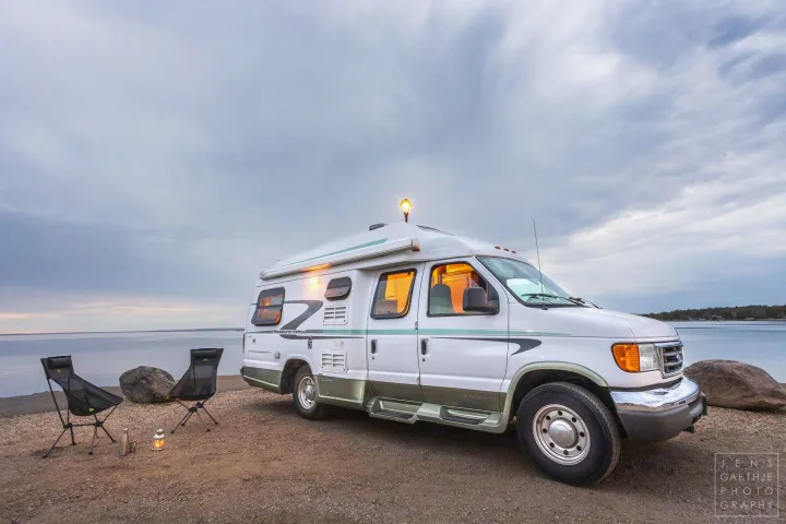 Cozy RV rental parked by a lake at dusk, chairs set up for a peaceful evening with warm interior lights.. Ford FordE350 2006