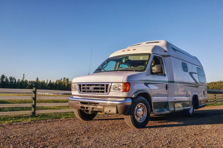 White Ford RV rental camper van parked on a gravel road. A fence and trees line the background under a clear blue sky.. Ford FordE350 2006