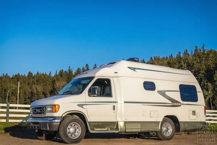 A white Pleasure-Way Excel-TS camper van for your next rv rental adventure, parked by a forest and fence under a clear sky.. Ford FordE350 2006