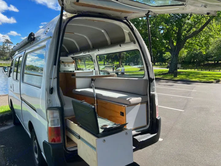 White rv rental campervan, rear open, showcases custom interior with kitchen pull-out & bench seating by a lake.. Nissan Caravan 2005