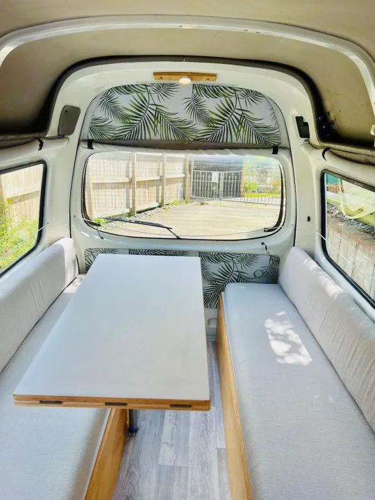 Bright, inviting RV rental interior: a white table, light grey cushioned benches, and tropical palm leaf accents on walls.. Nissan Caravan 2005