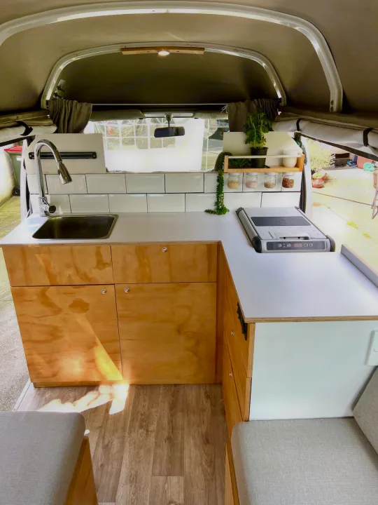 Inside an RV rental: a modern, compact kitchen with light wood cabinets, white counters, sink, and cooktop.. Nissan Caravan 2005