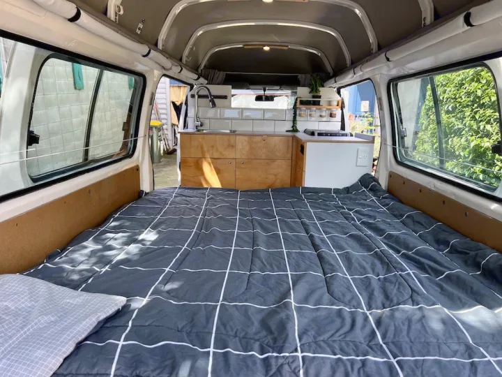 Inside a modern RV rental: comfy bed with grid comforter, full kitchen setup & natural light.. Nissan Caravan 2005