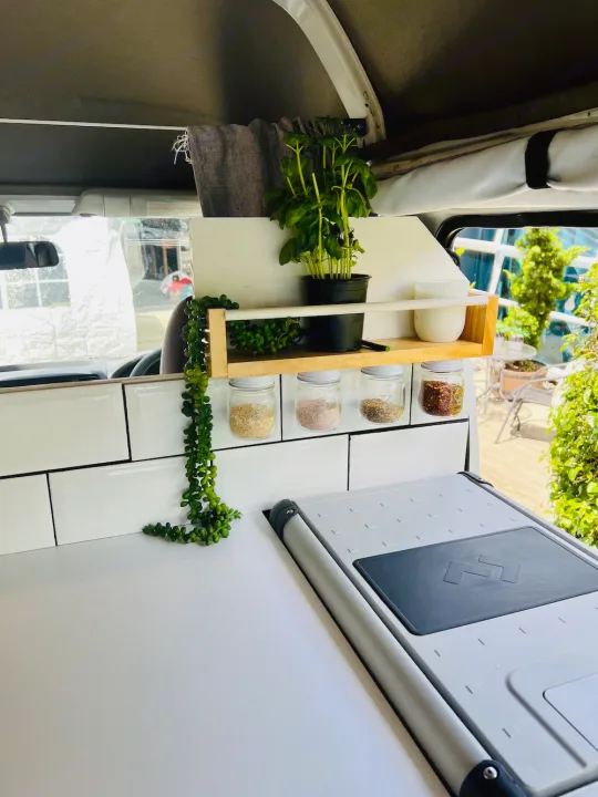 A cozy RV rental kitchen with white subway tile backsplash, organized spice jars, and a potted basil plant on a shelf.. Nissan Caravan 2005