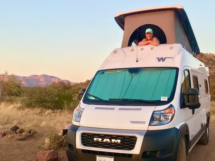 Woman enjoys the view from an RV rental pop-top camper van at a scenic mountain campsite.. Dodge Winnebago 2021