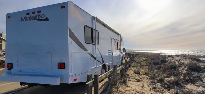 White Majestic RV rental parked on a sandy coastline next to a wooden fence, with the ocean sparkling under a cloudy sky.. Thor Motor Coach Four Winds Majestic 2017