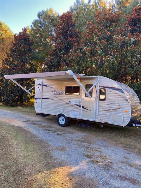 This beige RV rental, with its awning extended, is parked on a gravel path, perfect for your next outdoor adventure.. Shasta Oasis 18BH 2018