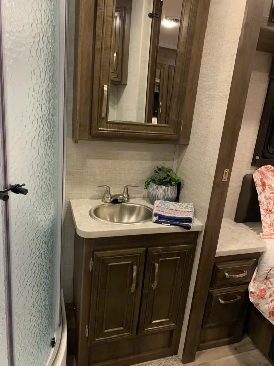 RV rental bathroom features a dark wood vanity with silver sink, mirrored cabinet, and textured shower door. Adjacent bed area visible.. Ford Jayco RedHawk 2021