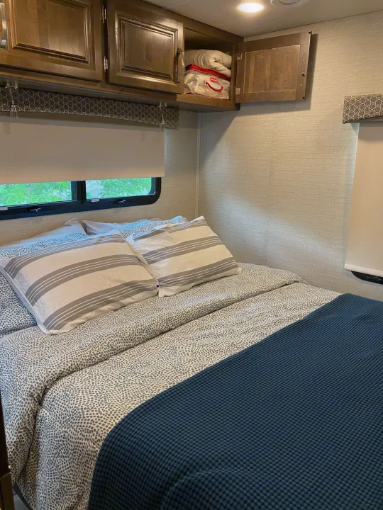 Cozy RV rental bedroom features a comfy bed with striped pillows, patterned bedding, open overhead cabinet, and windows.. Jayco Greyhawk 2019