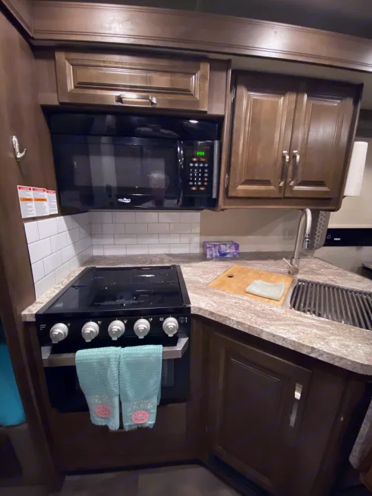 Detailed view of an RV rental kitchen: microwave over a stove, L-shaped counter with a sink, and dark wood cabinets.. Jayco Greyhawk 2019