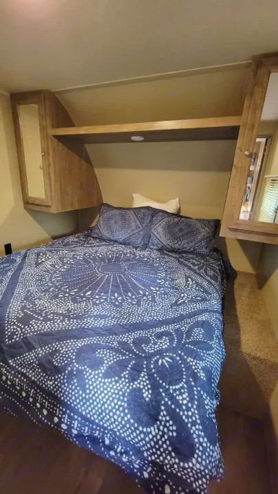 Cozy RV rental bedroom featuring blue patterned bedding, a white pillow, built-in wooden storage, and an overhead mirror.. Forest River Puma 2017