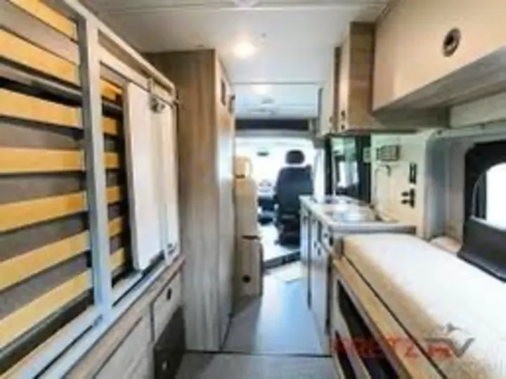 View inside an RV rental: a narrow aisle, compact kitchen, bed, and driver's area beyond. Modern camper van interior for travel.. Winnebago Solis 59PX 2022