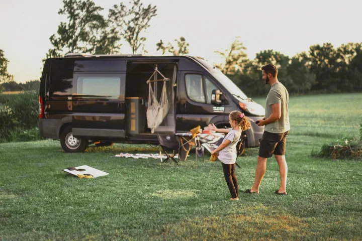 RV rental family fun: A dad and daughter play games in a grassy field next to their open camper van and camp setup.. Other Other 2015