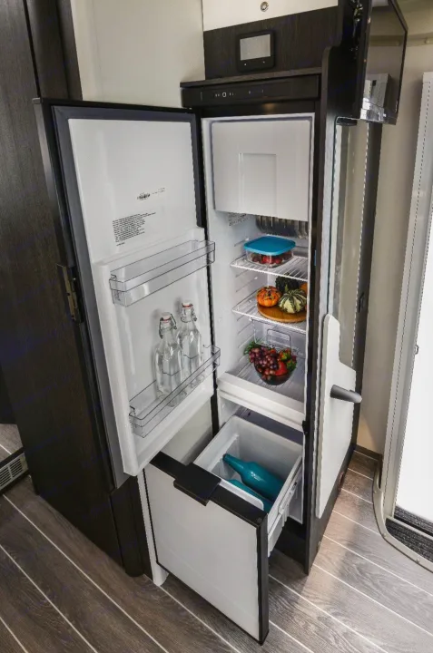 Open RV rental refrigerator, revealing fresh produce, bottles, and a pull-out drawer inside a compact RV kitchen.. Roller Team 685 Zefiro 2022