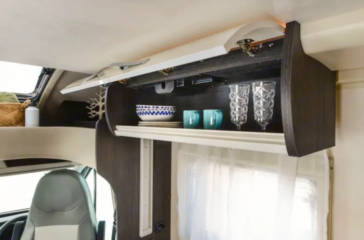 Overhead cabinet storage in an RV rental, neatly stocked with dishes, cups, and glasses, beside a window with sheer curtains.. Roller Team 685 Zefiro 2022