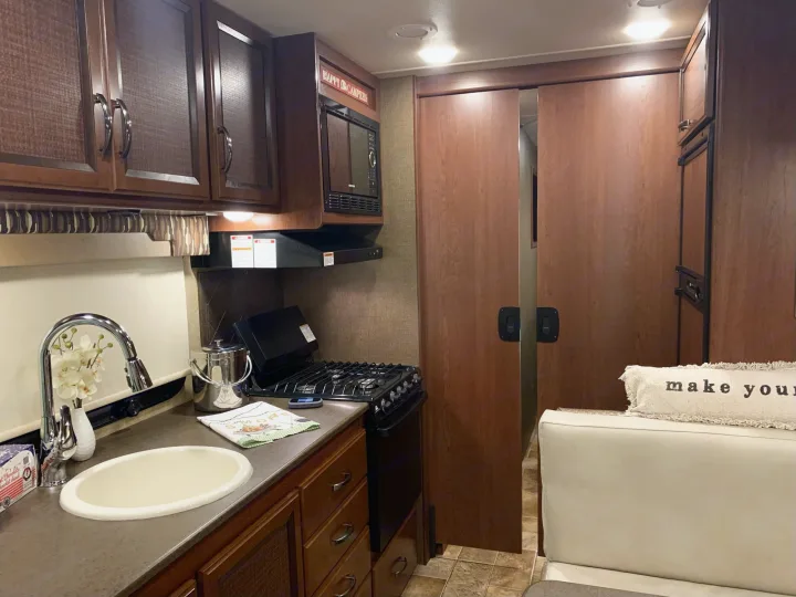 An RV rental kitchen features a sink, stove, microwave, and wood cabinets. Sliding doors reveal another interior space.. Thor Vegas 2016