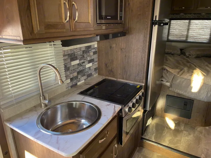 RV rental interior: A compact kitchen with a sink, stove, microwave, wood cabinets, and fridge. Part of a bed is visible.. Gulf Stream Conquest 2021