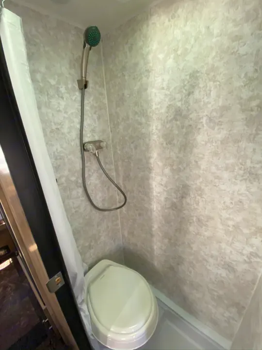 Compact RV rental wet bath with a toilet and handheld shower, featuring light gray textured walls and a white curtain.. ROULOTTE PROLITE ROULOTTE PROLITE 2020