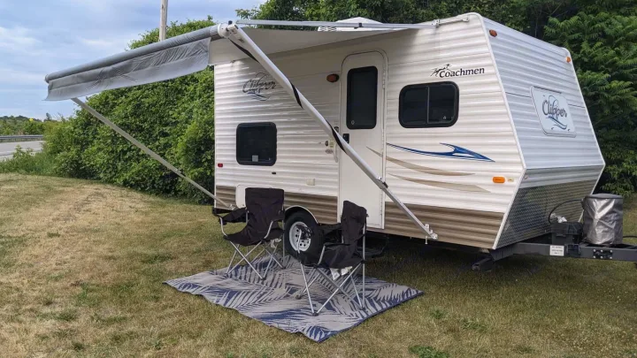 This RV rental travel trailer is set up for outdoor relaxation with an extended awning, two chairs, and a rug on the grass.. Coachmen Clipper 2013