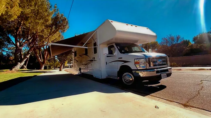White RV rental with its awning extended, parked on a sunny paved area surrounded by large trees.. Winnebago 31k 2015