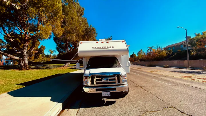 White Winnebago **RV rental** parked on a sunny day next to a green park with trees and a street.. Winnebago 31k 2015