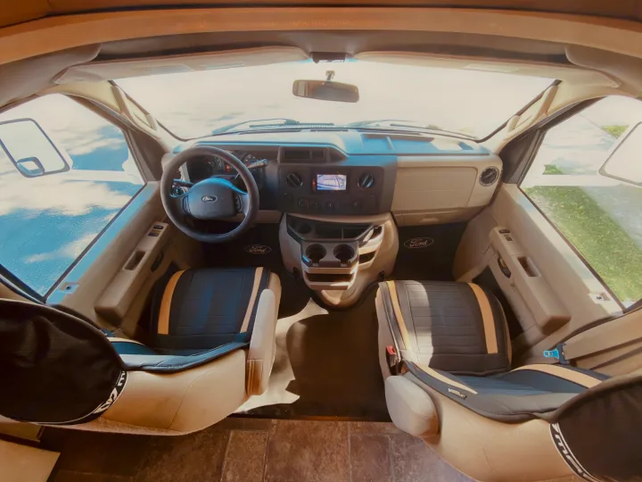 View of an RV rental's bright front cabin, showing Ford dashboard, steering wheel, and two custom-covered seats.. Winnebago 31k 2015