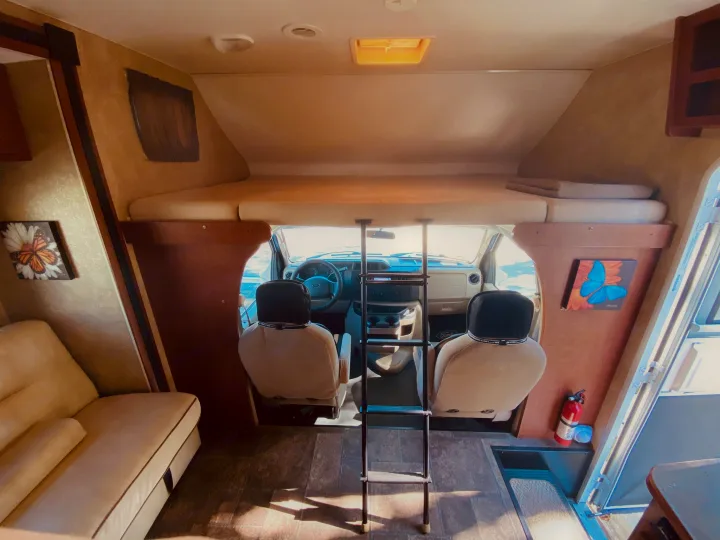Bright RV rental interior: overhead bed with ladder, cockpit, and lounge seating. Fire extinguisher on the right.. Winnebago 31k 2015