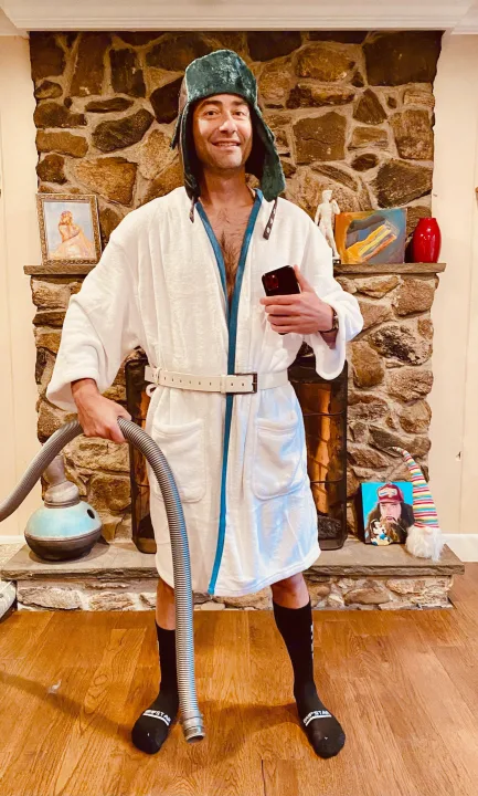 Man in Clark Griswold robe & hat, with a vacuum hose, ready for an RV rental journey. Standing by a stone fireplace.. Thor Freedom Elite 2021