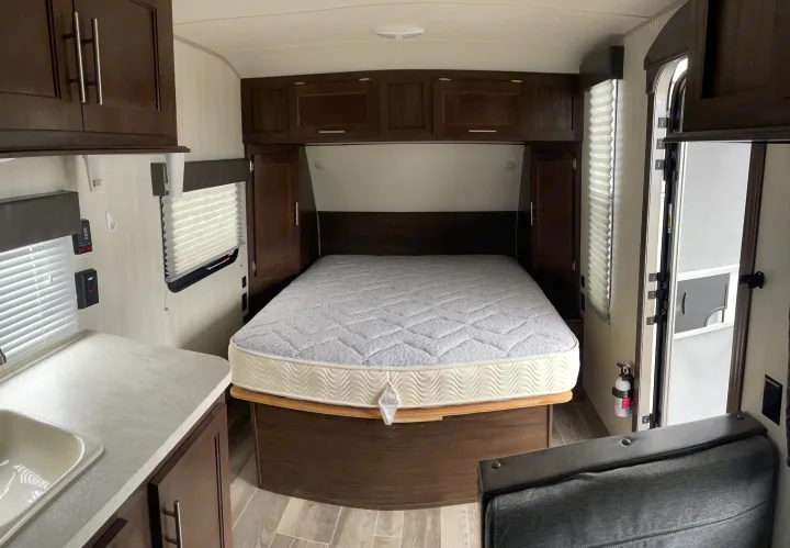 Inside an RV rental: a cozy bedroom with a queen mattress, dark wood cabinets, and a glimpse of the kitchen sink and living area.. Forest River Cherokee Wolf Pup 2019