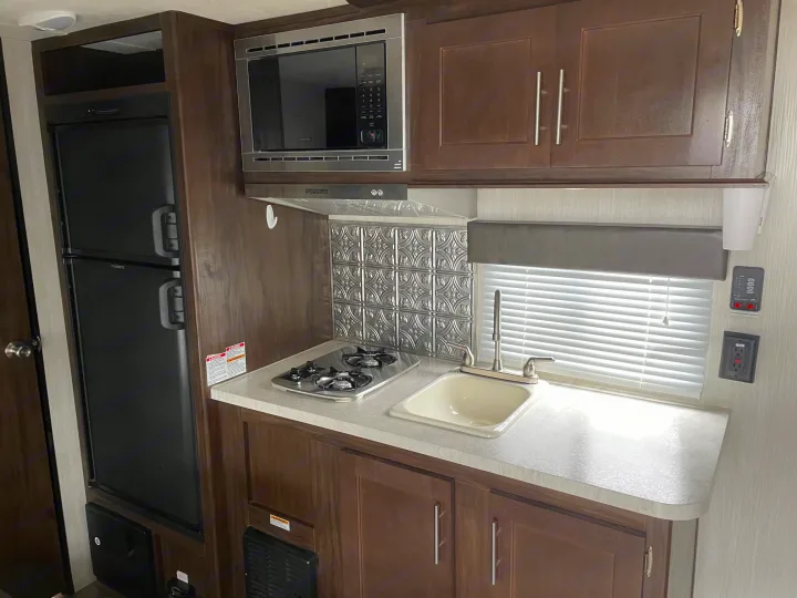 Compact RV rental kitchen: black refrigerator, stainless microwave, 2-burner stovetop, sink, and wood cabinets.. Forest River Cherokee Wolf Pup 2019