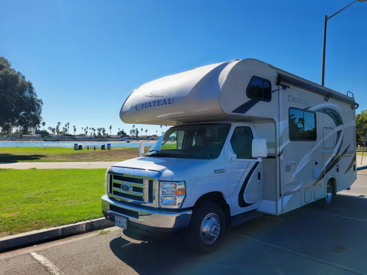 A Chateau rv rental is parked in a sunny park beside a bay, with lush grass and palm trees under a clear sky.. Thor Chateau 2018