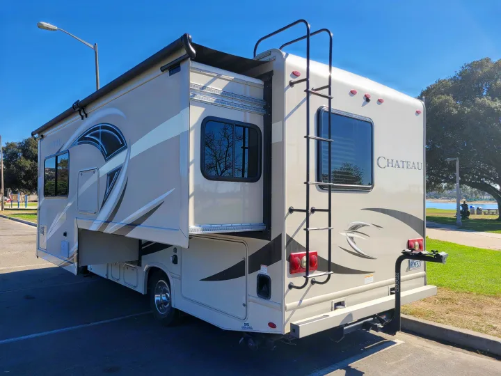 Experience outdoor adventure! This beige RV rental with a slide-out and bike rack is parked by water and trees under a clear sky.. Thor Chateau 2018