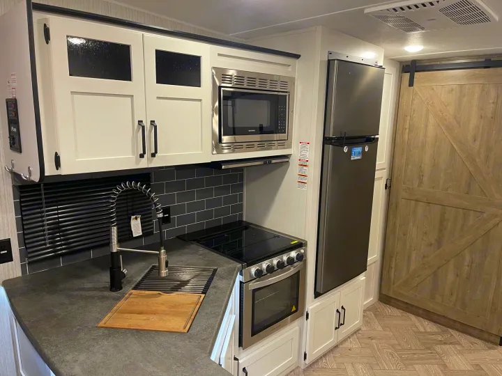 Modern RV rental kitchen with white cabinets, stainless steel appliances, dark counters, and a barn door. Great for cooking!. Palomino Puma 2022